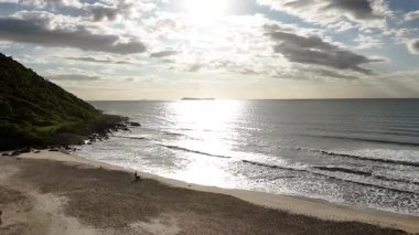 Santa Catarina, Brezilya 'da kıyıya vuran dalgalar, kıyıda yeşil bir yamaç ve okyanusa yansıyan günbatımının hava aracı görüntüleri. Ufukta görünen adaların olduğu manzara seyahat videosu.