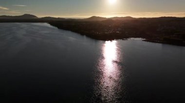 Santa Catarina, Brezilya 'daki bir göl üzerindeki hava aracı görüntüleri. Güneş dağların ardında batıyor ve suya yansıyor. Doğal manzarası ve şehir silueti olan manzaralı seyahat videosu..