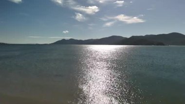 Brezilya, Santa Catarina 'daki sakin okyanusun üzerinde ilerleyen hava aracı görüntüleri. Videoda güneş ışığı yansıması, ufuktaki uzak dağlar ve huzurlu bir kıyı manzarası olan köpüklü su gösteriliyor..