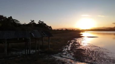 Sakin bir gölün üzerinde güzel bir günbatımının yumuşak ve huzurlu bir görüntüsü. Video, kıyı şeridi ve eski yapıların görüntüsüyle başlıyor, sonra suyun üzerinde pürüzsüz bir şekilde dönüyor, güneşin altın ışığının çarpıcı bir yansımasını ortaya çıkarıyor..
