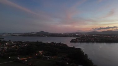 Büyüleyici bir dron atışı, Brezilya 'daki bir lagünün üzerinde alacakaranlığın sakin güzelliğini yakalar. Gökyüzü canlı günbatımı renkleriyle parlıyor, dağların ve ağaçların siluetini aydınlatıyor, şehrin ilk ışıkları kıyıda parıldamaya başlıyor..