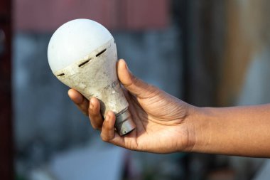 Close-up of a female hand holding a dirty, used LED light bulb against a blurred background. The old, bad bulb is a symbol of energy waste and obsolescence.