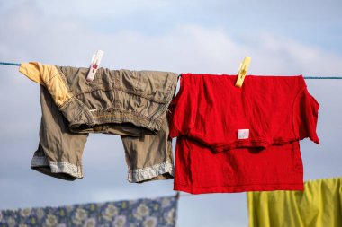 Canlı kırmızı bir tişört ve pantolon yakın plan çamaşır ipinde kuruyor, güneşli bir çatıda mandallarla güvence altına alınmış, parlak mavi bir gökyüzüne karşı..