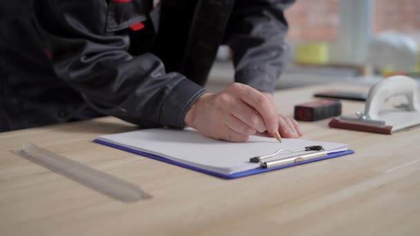 Il costruttore sta disegnando un progetto su un foglio di carta usando la matita, primo piano delle mani su un tavolo — Video Stock