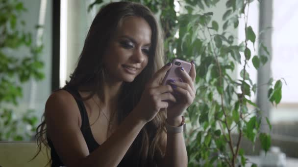 Очаровательная брюнетка девушка печатает на сенсорном экране своего смартфона, сидя в ресторане в дневное время — стоковое видео