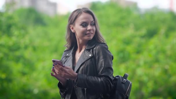 Jeune femme est debout à l'extérieur dans la journée ensoleillée et en utilisant son téléphone mobile, composer le numéro et regarder autour — Video