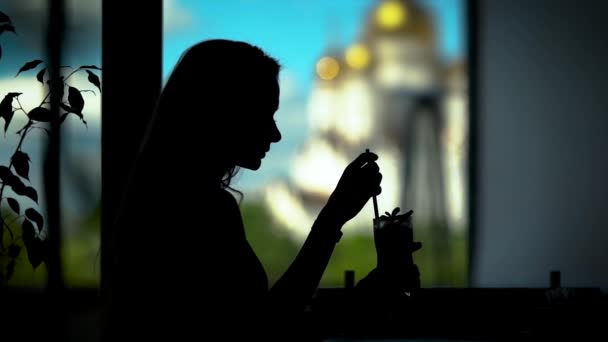 Черный силуэт молодой женщины, пьющей коктейль из стекла через солому в кафе, напротив окна — стоковое видео