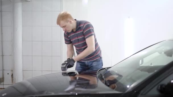 Rothaarige männliche Arbeitskraft bearbeitet die Oberfläche des Autos mit einer Poliermaschine in einem Autoservice und deckt sie mit einer Schutzschicht ab — Stockvideo