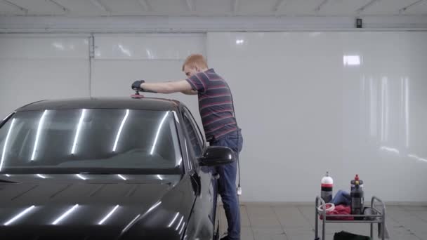 Redhead man is using polishing machine for smoothing surface of car, touching by tool a roof of auto in a garage — Stock Video