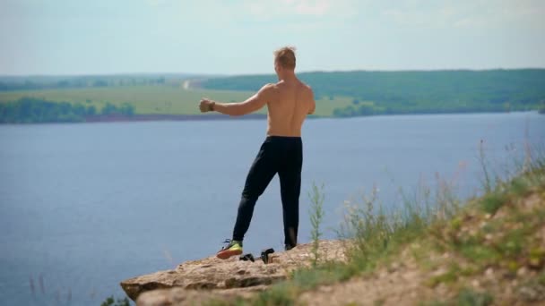 Deportista con el torso desnudo está llegando al borde de la montaña en el día soleado y levantando las manos, regocijando algo — Vídeos de Stock