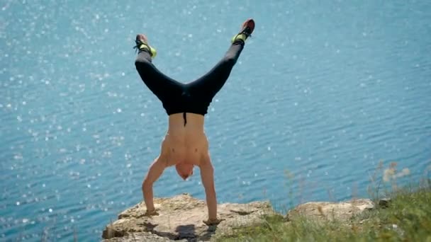 Sportlicher Mann steht an einem sonnigen Sommertag kopfüber über der Klippe, im Hintergrund blaues Meer — Stockvideo