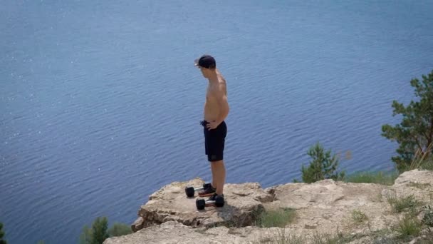 Young powerlifter is lifting dumbbells by both hands standing on edge of cliff over sea in summer day — Stock Video