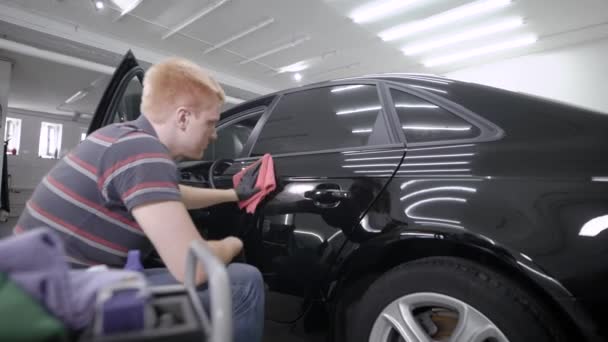 Працівник витирає двері розкішного чорного автомобіля після захисної обробки і миття в гаражі, сидячи поруч — стокове відео