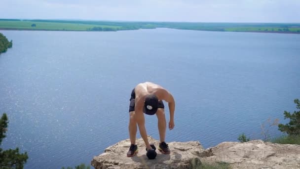 Jovem powerlifter está levantando halteres por ambas as mãos de pé na borda do penhasco sobre o mar no dia de verão — Vídeo de Stock