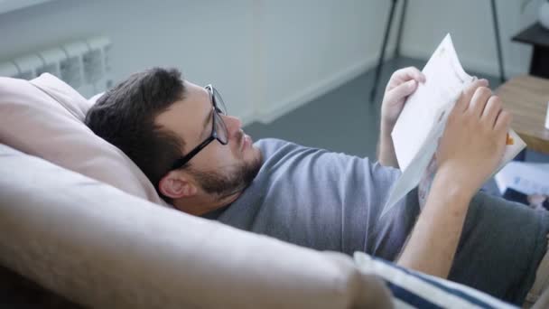 Porträt eines Mannes, der zu Hause auf dem Sofa eine Zeitschrift liest — Stockvideo