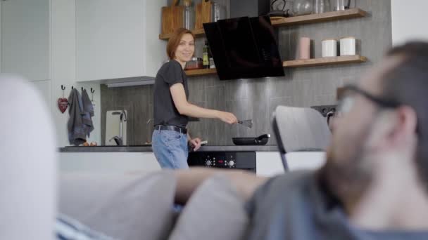 Il marito si siede sul devano, mentre sua moglie sta preparando la colazione. I giovani vivono insieme — Video Stock