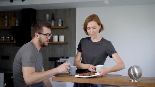 Χαριτωμένο νεαρό ζευγάρι πίνοντας τον καφέ στην κουζίνα. Σύζυγος και η σύζυγος κάθονται δίπλα-δίπλα σε ένα μοντέρνο εσωτερικό. — Αρχείο Βίντεο