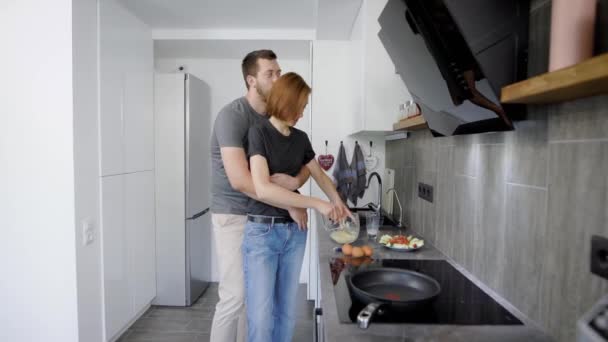 Kvinnan matlagning frukost i köket medan hennes make kramar henne bakifrån. — Stockvideo