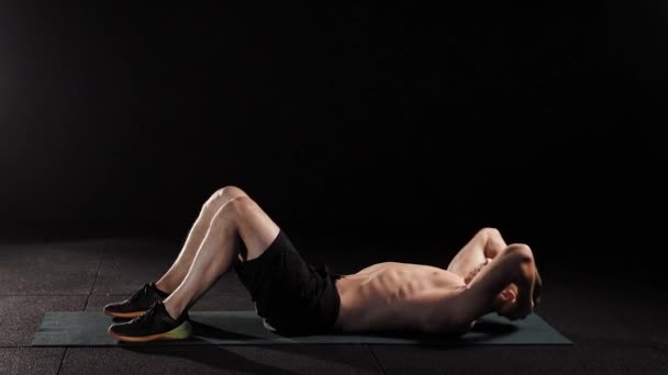 L'homme athlétique travaille les muscles de l'abdomen, couché sur le sol dans la pièce sombre de la salle de gym, soulevant le torse, tenant la tête par les mains — Video