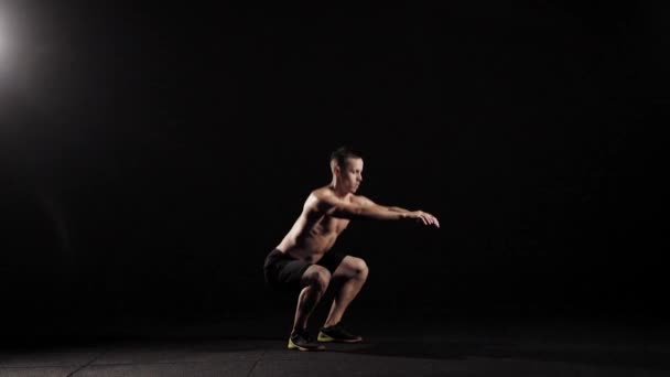Sterke jongeman met naakte torso doet gehurkt in een fitness zaal in avond, stretching handen vooruit — Stockvideo