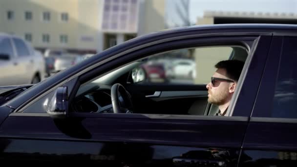 Un joven con barba cabalga en el estacionamiento en su coche negro. sedán de negocios se mueve tranquilamente a lo largo de otras máquinas . — Vídeos de Stock