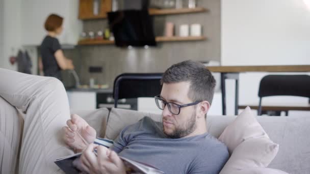 Mann ruht sich am Sonntagmorgen auf Couch mit Magazin aus, seine junge Frau kocht mit Herd in Küche — Stockvideo