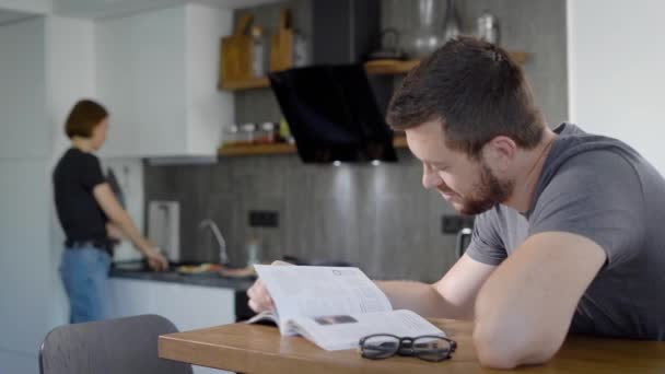 Mulher está cozinhando café da manhã para seu amado marido de manhã, o homem está sentado à mesa e livro de leitura — Vídeo de Stock