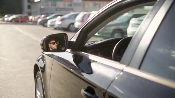 Άνδρας οδηγεί μεγάλο αυτοκίνητο σε δρόμους της πόλης, θέα από το εξωτερικό, το πρόσωπό του αντικατοπτρίζοντας σε ένα καθρέφτη σε ηλιόλουστη μέρα — Αρχείο Βίντεο