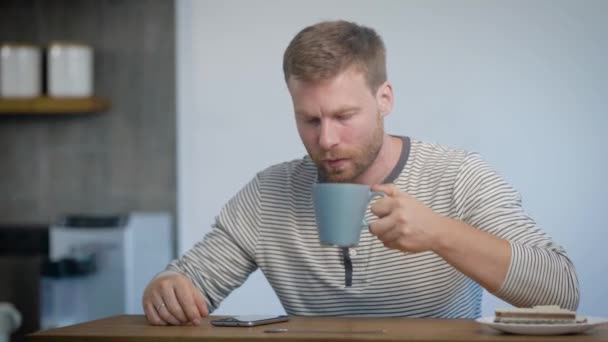 Sozinho jovem está tomando café da manhã em seu apartamento, sentado na cozinha, bebendo café e olhando em uma exibição de smartphone — Vídeo de Stock