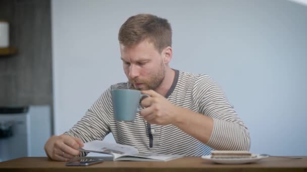 Jovem com barba bebendo café e lendo revista na cozinha pela manhã . — Vídeo de Stock