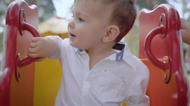 Niedliche kleine Junge mit großen Augen sitzt auf Kinderschaukel auf Spielplatz im Sommer Tag, Blick in die Kamera — Stockvideo
