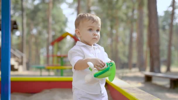 Funny baby boy is standing in sandbox in summer day in park, playing with plastic toy alone — Stock Video