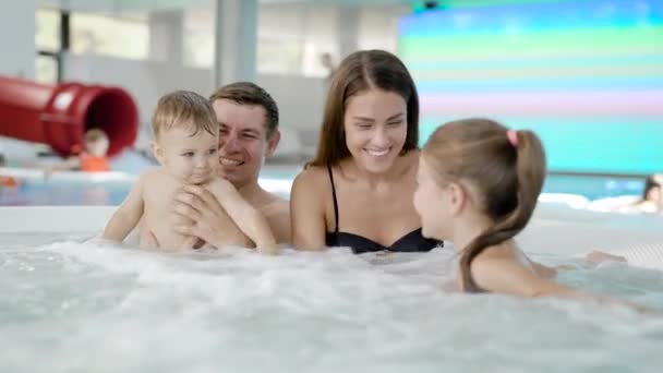 Famille heureuse avec deux petits enfants nagent dans une piscine, assis dans un petit bol de jacuzzi pulvérisation — Video