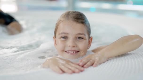 Menina bonita com cabelo molhado está sentado sozinho no banho de hidromassagem em um parque aquático, olhando para a câmera — Vídeo de Stock
