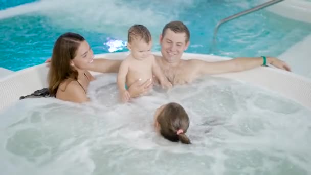 Счастливые родители отдыхают в аквапарке со своими детьми, лежа в чаше джакузи, мать держит мальчика — стоковое видео
