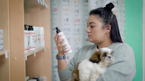 Young female pet owner is choosing shampoo for her dog in a shop, holding cute shih tzu puppy on hands — Stock Video