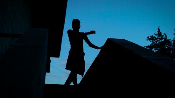 Dramatische silhouet van een jonge man is op een dak van huis dansen in de nacht tegen de donker blauwe hemel — Stockvideo