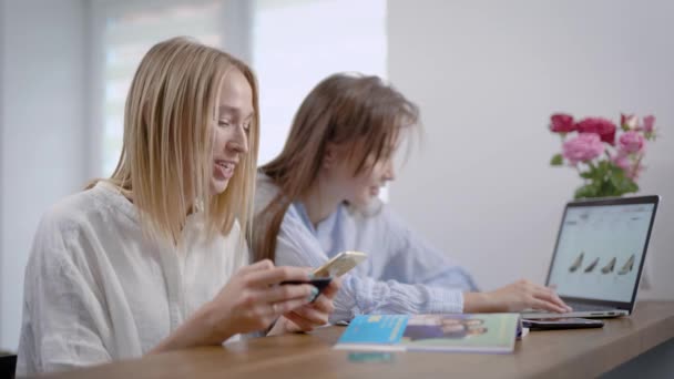 Две молодые девушки друзья покупают обувь в интернет-магазине, одна женщина платит с помощью кредитной карты, используя приложение на своем смартфоне — стоковое видео