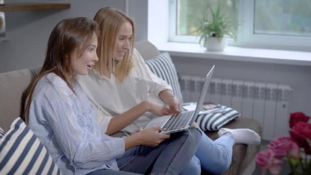 一对朋友的女孩坐在沙发上在家里和网上购物, 讨论和持有笔记本电脑上圈 — 图库视频影像