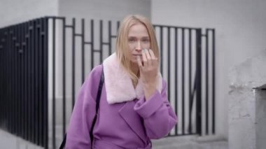 Sevimli sarışın kadın ile çit building yakınındaki şehirde yürüyüş kürk yakalı trendy leylak rengi ceket giyiyor