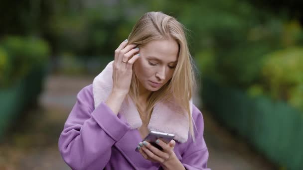 Adult blonde businesswoman is standing alone in park and typing message on her smartphone in daytime — Stock Video