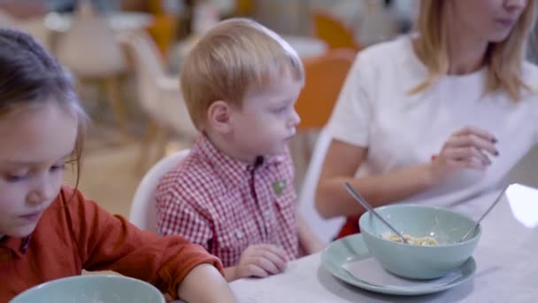 Mooie jonge moeder in een café. Een vrouw voedt haar zoon met pasta, naast haar dochter eet haar eigen schotel. — Stockvideo