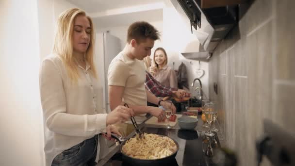 Unga människor matlagning tillsammans i moderna kök, två kvinnor tvättar grönsaker, andra flicka omrörning pasta — Stockvideo