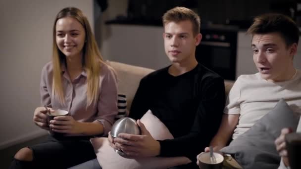 Cinco amigos estão assistindo filmes por tv sentados em um sofá na sala de estar na noite de férias, bebendo chá e café — Vídeo de Stock