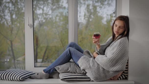 Mooie jonge vrouw zit op windwosill met glas rode wijn, warme gezellige weekenddag thuis. — Stockvideo