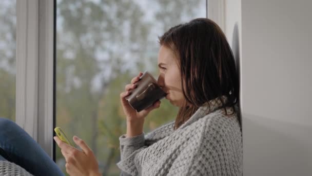 Linda chica en ropa de abrigo sentado en el alféizar de la ventana con un teléfono inteligente y una taza de té caliente, tiempo libre en fin de semana . — Vídeos de Stock