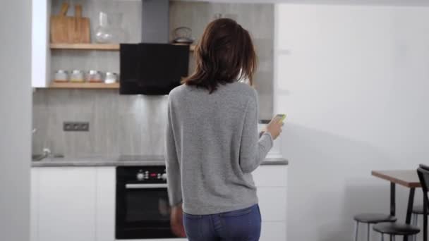 Ung kvinna med kort hår promenader i köket, lägger telefonen på bordet och börjar läsa en bok. — Stockvideo