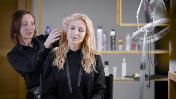 Charming blone girl sitting in chair of a beauty salon and looking forward whle stylist working with her long blonde hair. — Stock Video