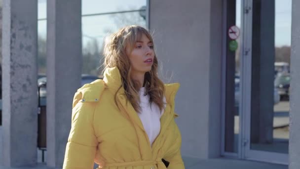 Young attractive blondie walking in city wearing stylish yellow coat in autumn, passing by a building. — Stock Video