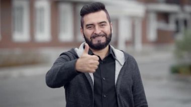 sakallı kendine güvenen adam açık havada gündüz de kameraya, gülümseyerek ve başparmak gösterilmesini seyir duruyor